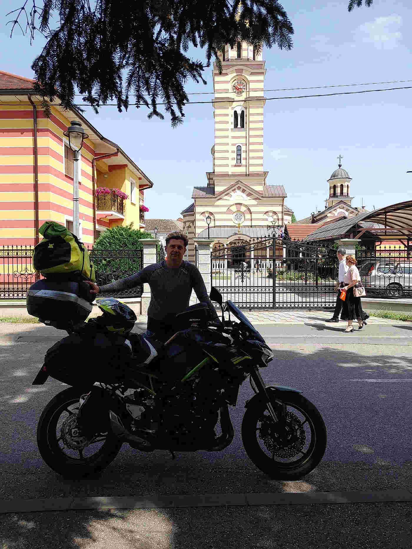 Motocyklista z wyprawy CARAVANA wyjazdy motocyklowe podczas podróży po Bośni i Hercegowinie, stojący przy zapakowanym motocyklu na tle charakterystycznego kościoła w bałkańskim miasteczku.