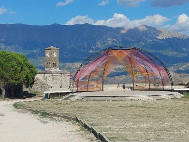 Wieża zegarowa i nowoczesna konstrukcja z kolorowym zadaszeniem podczas wyprawy motocyklowej Albania – Macedonia, bałkański krajobraz i góry.